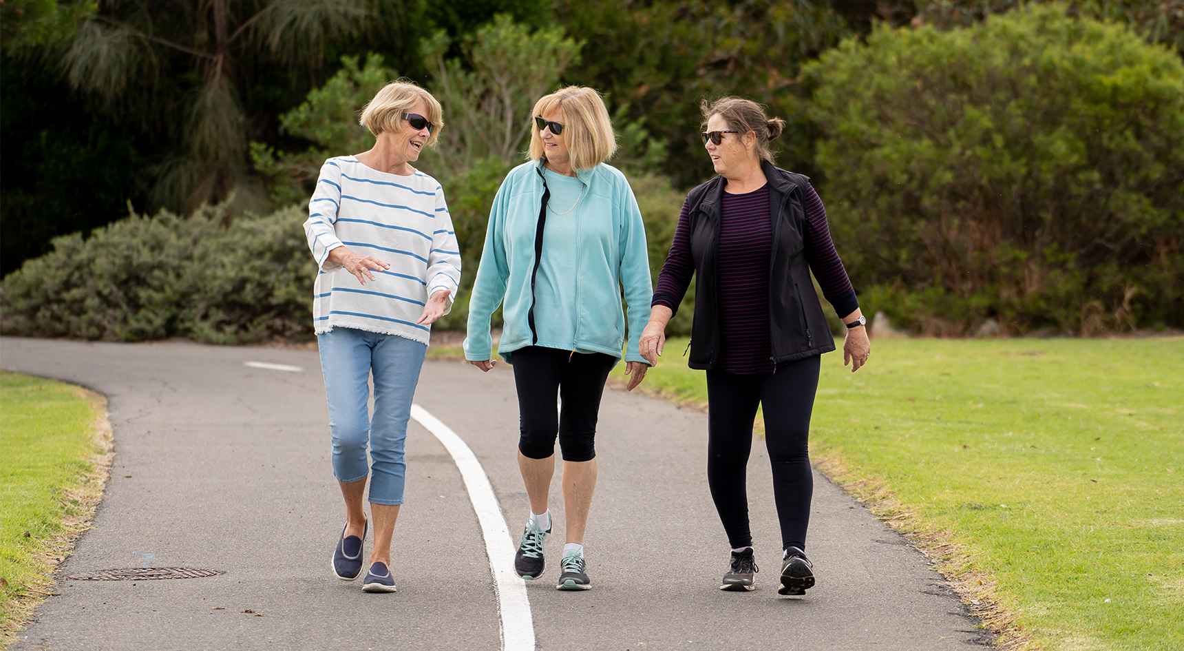 Three women in their 60s walking on a track through a park
