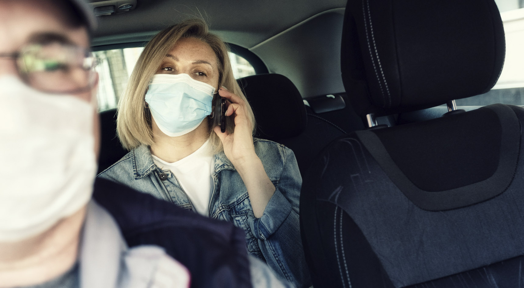Lady sitting in the back seat of a rideshare vehicle. Driver in front. Both people are wearing masks.