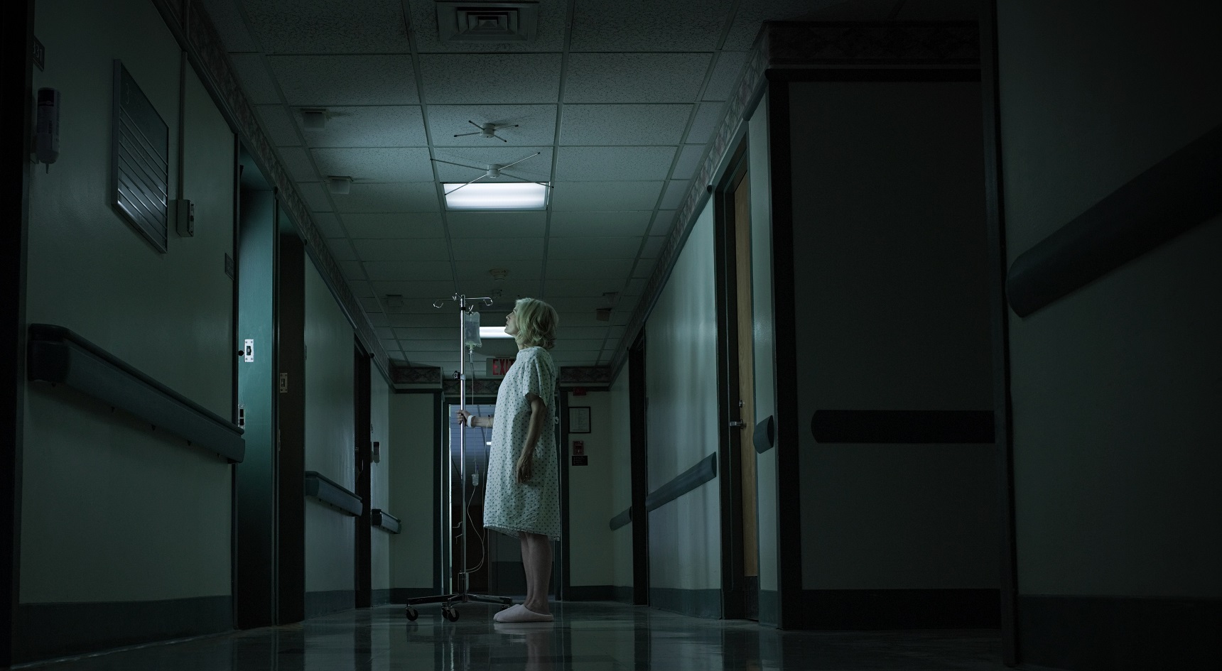 Female patient with intravenous drip stands in dark hospital corridor.