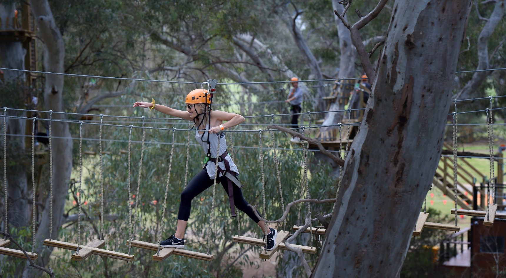 Woman with orange helmet crossing a wooden bridge among the treetops.a