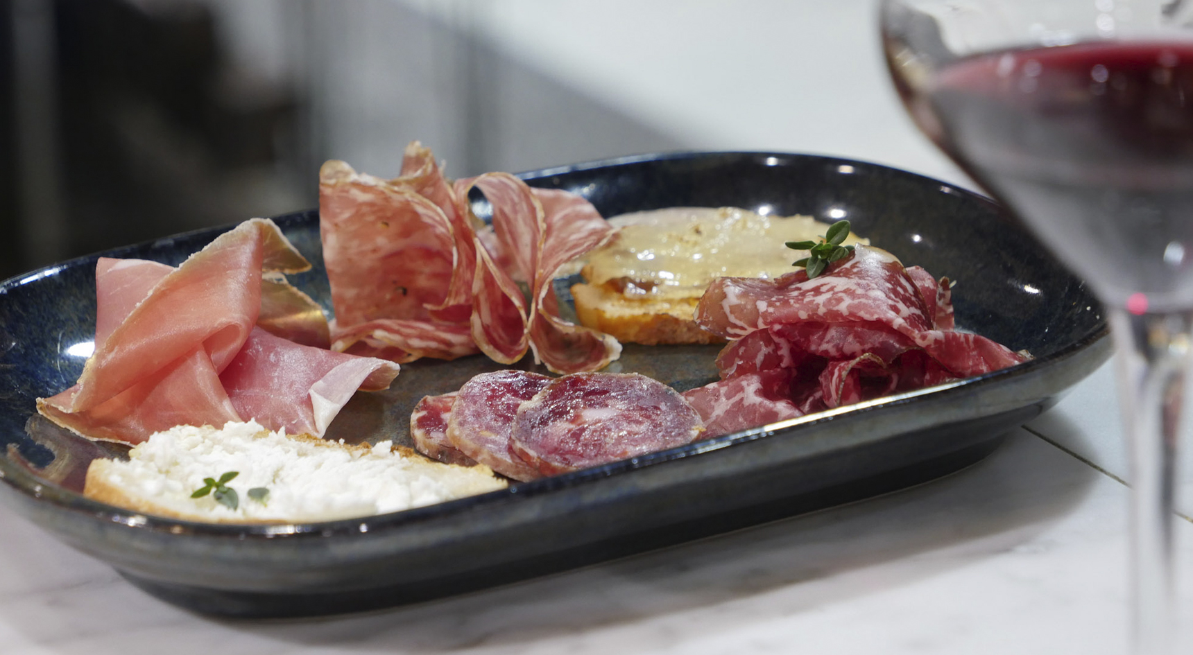 A variety of cured meats on a plate with bread and a wine glass to the side of the image.