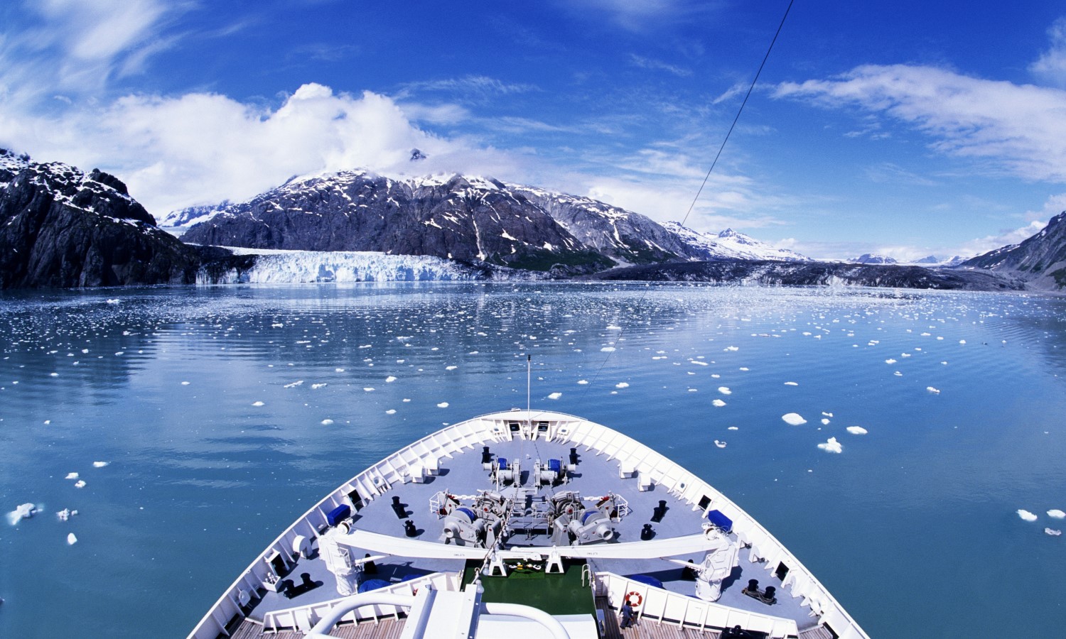 Front of a cruise ship on blue water with black and snow capped mountains.