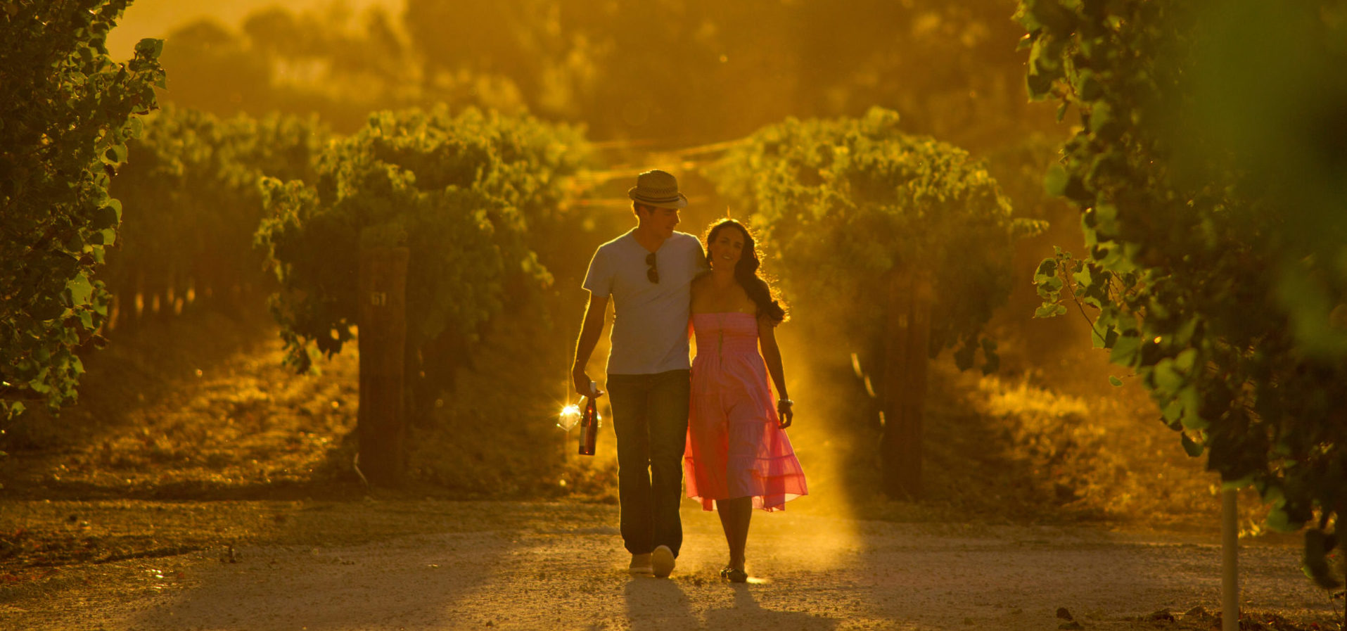 Couple walking through a vineyard with a bottle of wine.
